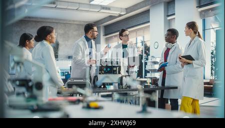 Diverse Team of Industrial Robotics Specialists Gathered Around a Table With a Mobile Robot. Engineers In Lab Coats Discussing an Automated AI Robotic Delivery Assistant, Using Laptop, Tablet Stock Photo