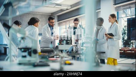 Diverse Team of Industrial Robotics Specialists Gathered Around a Table With a Mobile Robot. Engineers In Lab Coats Discussing an Automated AI Robotic Delivery Assistant, Using Laptop and Tablet Stock Photo