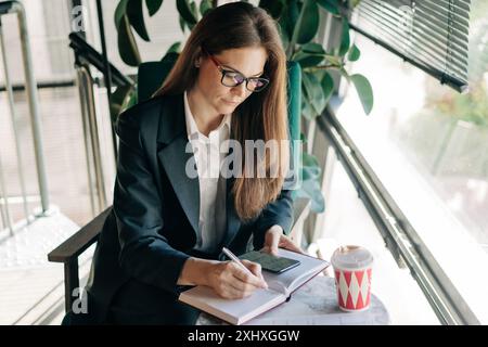 Business woman investor writes data from a mobile application into a notepad while sitting in a cafe. Stock Photo