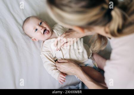 Mom changes the bodysuit of a cheerful baby after changing the diaper. Stock Photo