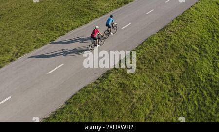 Side view of a Caucasian man and woman, in cycling wear, training for a race, riding road bikes in the middle of the paved road, aerial tracking drone Stock Photo