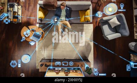 Top View Of Caucasian Man In the Loft Apartment Sitting Down on The Couch and Connecting Smartphone to Convenient Smart Home System. VFX Edit Visualizing Connected Devices. Laptop, TV, Speaker. Stock Photo