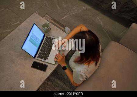 An overhead view of a woman working on her laptop, with a coffee cup and phone on the table. Cozy indoor setting. Stock Photo