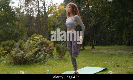 Caucasian sporty woman standing on one leg rotate hip warms up hips joints bend knee back sport stretching balance stretch workout yoga in park Stock Photo