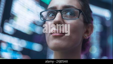 Close Up Portrait of Smiling Woman Looking Around, Lines of Code Language Reflected on her Glasses from Big Display Screens Surrounding her. Professional Female Programmer Developing Software, Coding Stock Photo