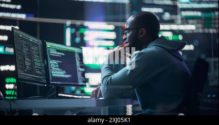 Focused Black Male Programmer Working in Monitoring Room, Surrounded by Big Screens Displaying Lines of Programming Language Code. Portrait of Man Creating Software. Abstract Futuristic Coding Concept Stock Photo