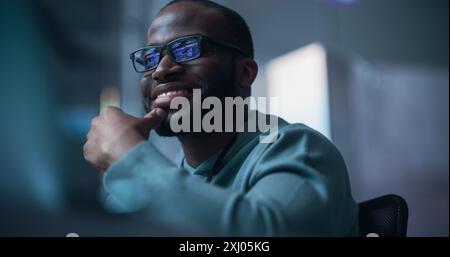 Close Up Of Black Male Developer Smiling Happily After Successfully Typing on Computer a Functioning Software Code. Professional Programmer Creating Mobile App, Screen Display Reflected on his Glasses Stock Photo