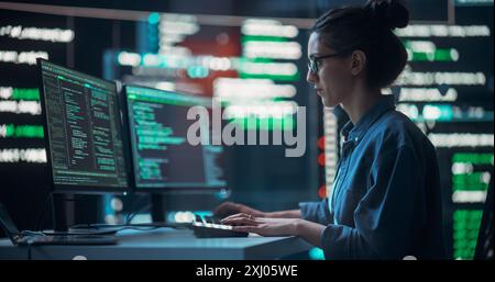 Medium Shot of Woman Working as a Developer, Surrounded by Big Screens Displaying Lines of Code in Dark Monitoring Room. Female Programmer Using Desktop Computer, Analysing Data, Creating AI Software Stock Photo