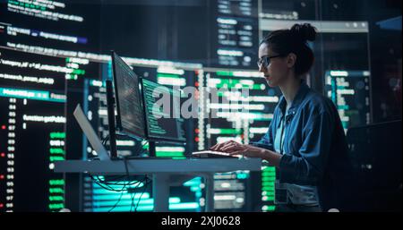 Portrait of a Woman Working on Computer, Typing Lines of Code that Appear on Big Screens Surrounding her in a Monitoring Room. Female Programmer Creating Innovative Software Using AI Data and System Stock Photo