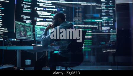 Portrait of a Black Man Working on Computer, Typing Lines of Code that Appear on Big Screens Surrounding him in Monitoring Room. Male Programmer Creating Innovative Software Using AI Data and System Stock Photo