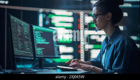Medium Shot of a Woman Working as a Developer, Surrounded by Big Screens Displaying Lines of Code in a Monitoring Room. Female Programmer Using Desktop Computer, Analysing Data, Creating AI Software Stock Photo