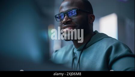 Black Male Developer Smiling Happily After Successfully Typing on Computer a Functioning Software Code. Professional Programmer Creating a Mobile App, Screen Display Reflected on his Glasses Stock Photo