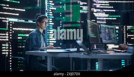 Portrait of a Woman Working on a Computer, Typing Lines of Code that Appear on Big Screens Surrounding her in a Monitoring Room. Female Programmer Creating Innovative Software Using AI Data and System Stock Photo