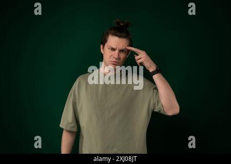 A man wearing a green shirt is standing with his hand pressed against his forehead. He appears to be deep in thought or experiencing some type of dist Stock Photo