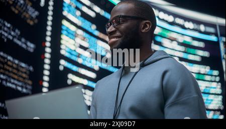 Portrait of Young Black Man Working on Laptop Computer, Looking at Big Digital Screen Displaying Back-end Code And Smiling. Professional Programmer Developing a Big Data Interface Software Project. Stock Photo