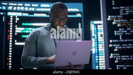 Portrait of Professional Programmer Developing a Big Data Interface Software Project.Young Black Man Working on Laptop Computer, Looking at Big Digital Screen Displaying Back-end Code Lines Stock Photo