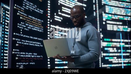Portrait of Young Black Man Working on Laptop, Looking at Big Digital Screen Displaying Back-end Code Lines. Professional Programmer Developing a Big Data Interface Software Project Concept. Stock Photo