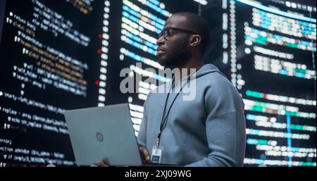 Portrait of Young Black Man Working on Laptop Computer, Looking at Big Digital Screen Displaying Back-end Code Lines. Professional Programmer Developing a Big Data Interface Software Project Concept. Stock Photo