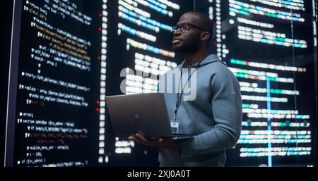 Portrait of Young Black Man Working on Laptop Computer, Looking at Big Digital Screen Displaying Back-end Code Lines. Professional Programmer Developing Big Data Interface Software Project. Stock Photo
