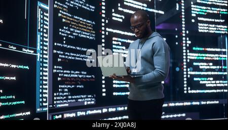Portrait of Young Black Man Working on Laptop Computer, Looking at Big Digital Screen Displaying Back-end Code Lines. Professional Programmer Developing a Big Data Interface Software Project Stock Photo
