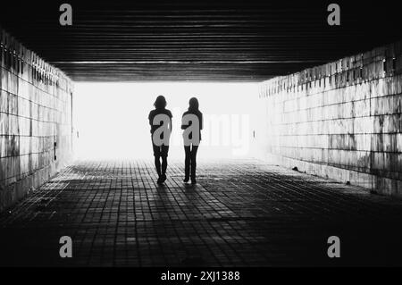 Silhouettes of two people in a tunnel. Black and white image with light coming from behind. Couple walking away from the camera. Stock Photo