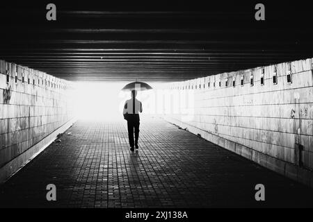 Silhouette of man with umbrella in a tunnel. Black and white image with light coming from behind. Person walking away from the camera. Stock Photo