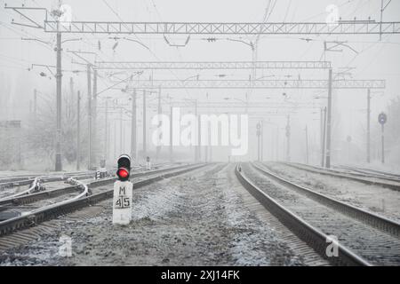 Railway tracks in a fog. Red signal light on railway semaphore. Low visibility - frost-covered railway tracks disappearing in a dense fog. Stock Photo