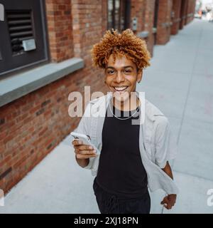 A young man with curly hair smiling and using his smartphone while walking down an urban street Stock Photo