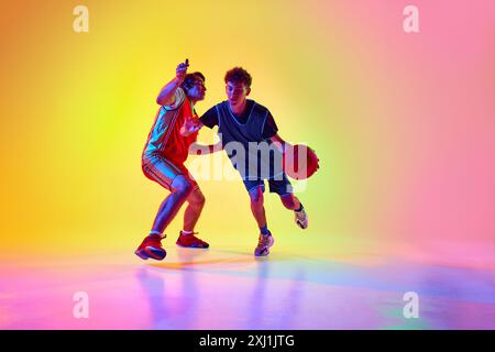 Two players, focused and energetic, engage in rigorous basketball training session in neon light against gradient studio background. Stock Photo