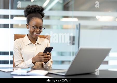 Confident woman smiling while using smartphone at office desk with laptop. Office environment with digital communication and workplace interaction. Stock Photo