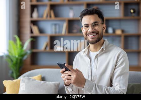 Smiling young man using smartphone in modern living room, seated on comfortable sofa. Bright and cozy home office setting, featuring wooden bookshelf and green plant background. Stock Photo