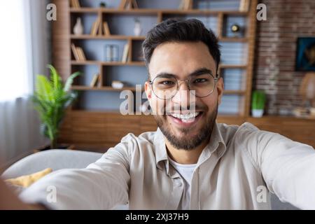 Close-up photo of young smiling Indian man in glasses and beard at home, holding camera and talking on video call. Stock Photo