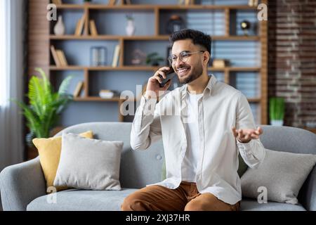 Smiling young Indian man in casual clothes and glasses talking on mobile phone gesturing with hands. Stock Photo