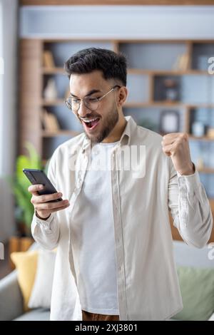 Young man feeling excitement while checking smartphone at home. Man with glasses and beard celebrating good news or achievement with fist pump and joyful expression Stock Photo