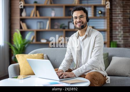 Portrait of smiling young Muslim man in glasses and headset sitting on sofa at table at home, using laptop, looking at camera. Stock Photo