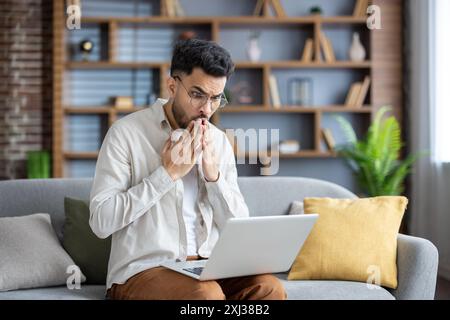 A shocked young Indian man sits on the sofa at home, holds a laptop on his lap, looks at the screen in surprise, covers his mouth with his hands in surprise. Stock Photo