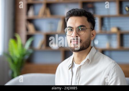 Close-up portrait of a young Indian man in casual clothes and glasses at home, sitting on the sofa and looking seriously at the camera. Stock Photo