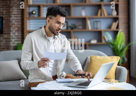 Focused young man working from home on couch analyzing financial documents using laptop. Remote work concept with home office setup displaying concentration and productivity Stock Photo