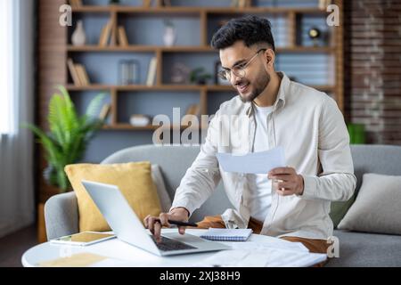 Indian young man working with documents and bills at home, sitting on sofa. holds papers and uses a laptop. Stock Photo