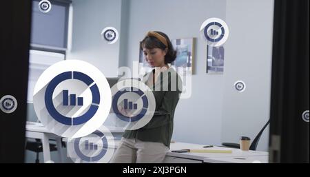 Image of multiple bar graph icons floating over asian woman using laptop at office. Computer interface and business data technology concept Stock Photo