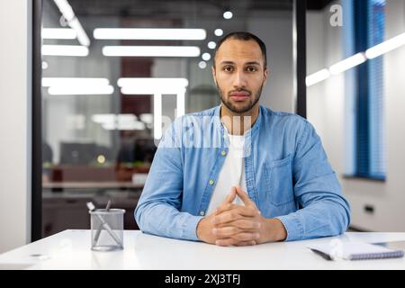 Portrait of young serious hispanic man sitting at desk in office, holding hands together and looking at camera. Stock Photo