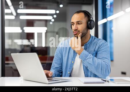 Thoughtful and serious Arab man, office worker, student working and studying in modern office wearing headphones and looking at laptop monitor screen. Stock Photo