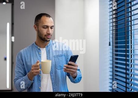 A young Hispanic man is standing by the window in the office lobby at work, holding a cup of coffee and using a mobile phone. Stock Photo