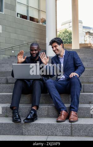 black African businessman and his Latin colleague sit on outdoor office stairs. They wave during a video call on a laptop, showcasing modern communica Stock Photo