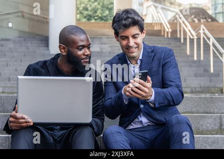 Hispanic businessman displays company data on smartphone to his African colleague, who holds a laptop, while they work outdoors on office stairs.highl Stock Photo