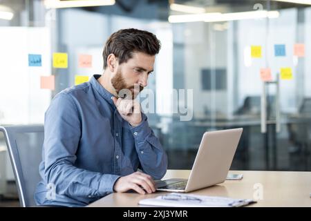 A serious and focused young man is sitting at a desk in the office and working on a laptop, looking thoughtfully at the screen. Stock Photo