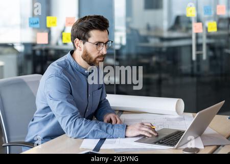 A serious and focused young man, an architect, surveyor and engineer, works in an office with desks and a laptop. Photo from the side. Stock Photo