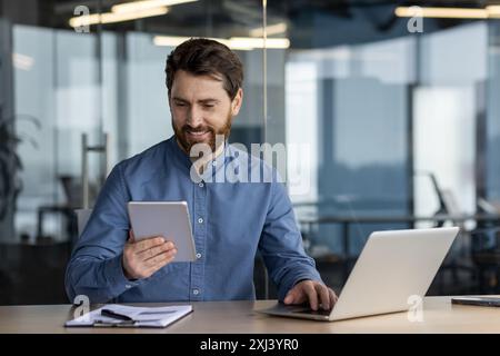A smiling young man works in the office at a notebook, holds in his hands and looks at the tablet screen. Stock Photo