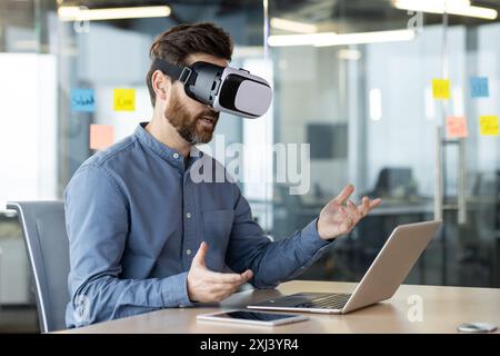 A young businessman works in the office, sits at a table with a laptop, wears a virtual mask and communicates online via video communication, gesturing with his hands. Stock Photo