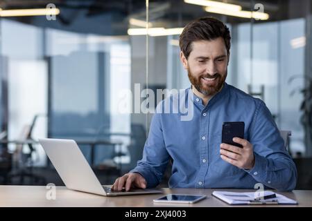 A smiling young man works in the office on a laptop, types on a keyboard and uses a mobile phone, talks on a video call. Stock Photo